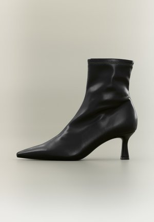 Bottine en cuir noir avec bout pointu et talon fin de hauteur moyenne, vue de profil sur fond clair uni.