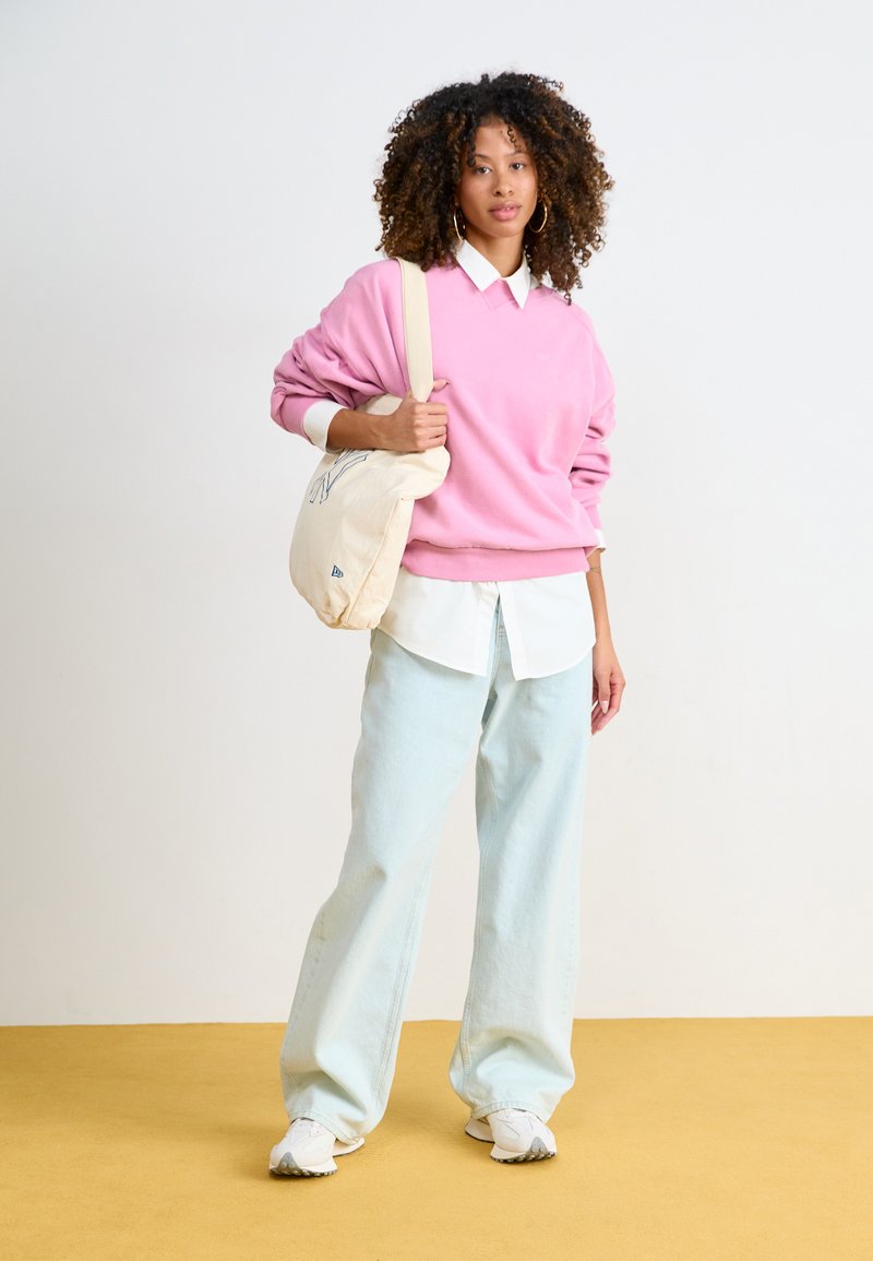 Rosa Sweatshirt, darunter ein weißes Hemd mit Kragen, hellblaue weite Jeans und weiße Sneaker. Hält eine cremefarbene Umhängetasche mit einem Logo.
