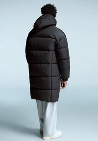 Svart dunjacka med huva, med quiltad design och lång modell, bärs över grå sweatpants och ljusa skor.