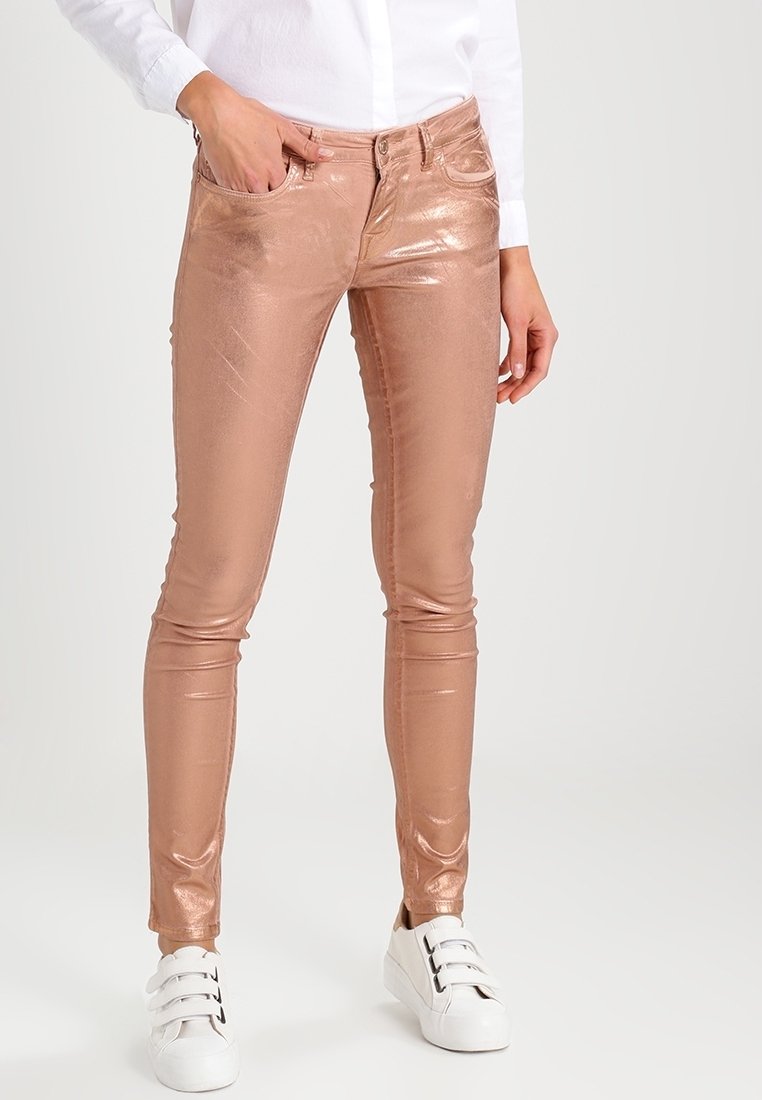 Pantalon slim en métal or rose porté avec une chemise blanche et des baskets blanches à scratch sur une personne debout.