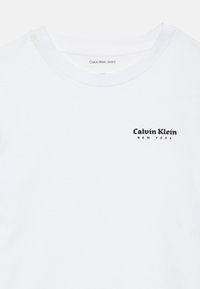 Valge T-särk musta "Calvin Klein New York" logoga trükituna vasakul rinnal krae all.