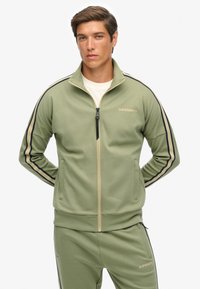 Groene trainingsjas met rits, voorzien van zwarte en beige strepen, een hoge kraag, ritssluiting en een logo op de borst. Glad stoffen textuur.