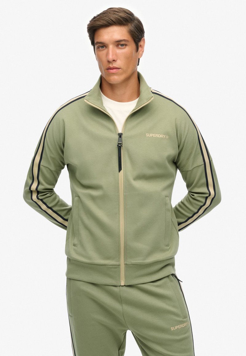 Groene trainingsjas met rits, voorzien van zwarte en beige strepen, een hoge kraag, ritssluiting en een logo op de borst. Glad stoffen textuur.