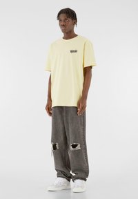 Yellow cotton t-shirt with short sleeves and black text on the chest, paired with gray distressed jeans and white sneakers.