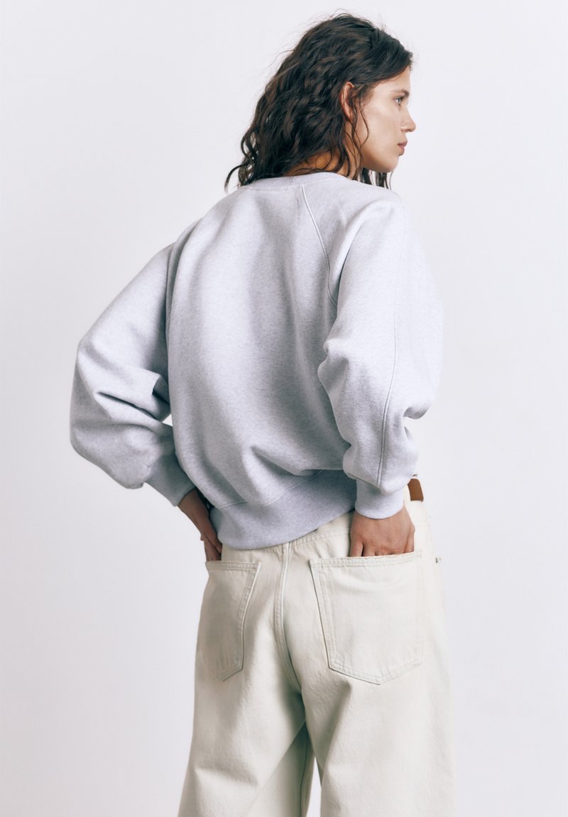 Kvinde med mørkt, bølget hår iført en lysegrå sweatshirt og beige bukser, med hænderne stukket i baglommerne, vendt sidelæns.