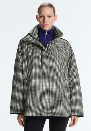 Žena v nosenej voľnej čierno-bielej károvanej bunde s vysokým golierom cez tmavomodrý top so zipsom na jednoduchom pozadí.