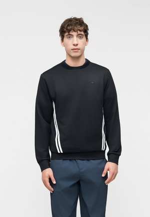 Junger Mann trägt einen schwarzen Pullover mit weißen vertikalen Streifen an den Seiten und blaue Hosen, steht vor einem schlichten Hintergrund.