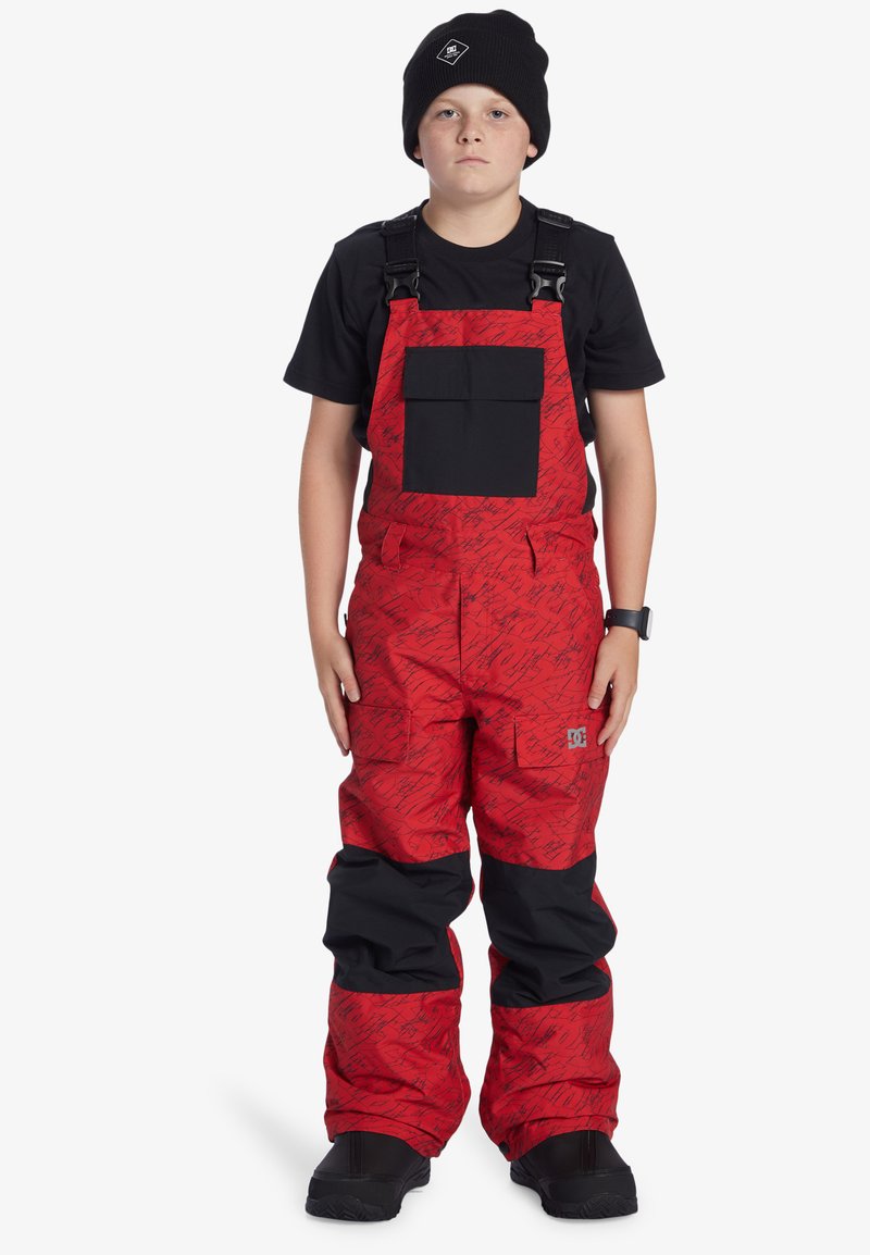 Röd snöbyxa med svarta detaljer, en stor bröstficka och ett stiliserat mönster; justerbara remmar och ankelfållar; bärs med en svart mössa.