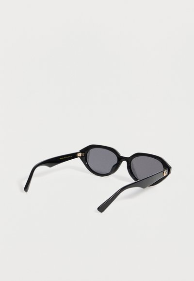Lunettes de soleil œil de chat noires avec verres teintés foncés et détails dorés aux charnières, posées sur une surface blanche.