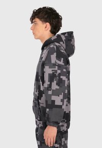 Młody mężczyzna w czarno-szarym kompozytowym kapturze i dopasowanych spodniach, stojący w profilu na tle jasnym.