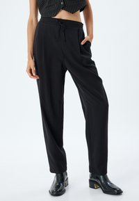 Pantalons noirs amples avec une taille élastique et un cordon de serrage. Possède des poches latérales et une texture lisse. Associés à des bottes noires à talons.