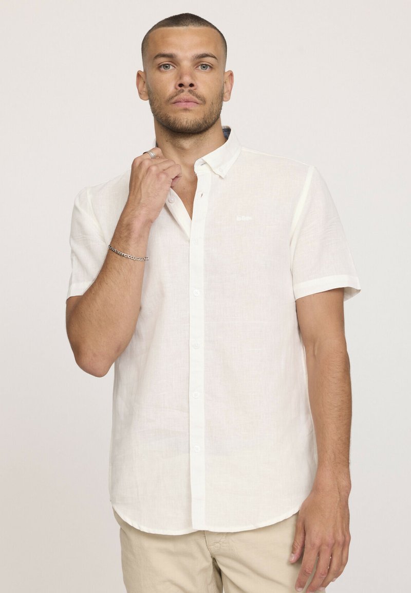 Chemise en lin blanche à manches courtes avec un col boutonné, présentant une texture subtile et un petit logo sur la poitrine.