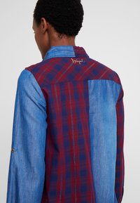 Persona che indossa una camicia a maniche lunghe con maniche in denim blu e schiena a quadri rossi e blu, con testo ricamato vicino al colletto.