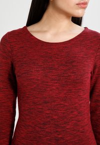 Un haut rouge à manches longues avec un col rond, présentant un motif texturé et chiné. Le tissu semble doux et léger.
