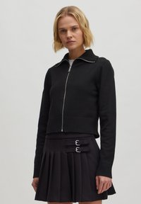 Schwarzer, kurze Pullover mit Reißverschluss und geripptem Kragen, kombiniert mit einem gefalteten schwarzen Rock mit zwei silbernen Schnallen.