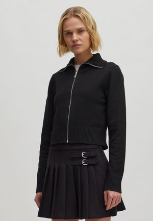Schwarzer, kurze Pullover mit Reißverschluss und geripptem Kragen, kombiniert mit einem gefalteten schwarzen Rock mit zwei silbernen Schnallen.