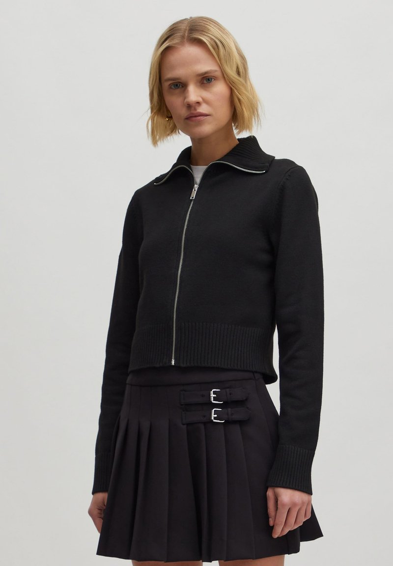 Schwarzer, kurze Pullover mit Reißverschluss und geripptem Kragen, kombiniert mit einem gefalteten schwarzen Rock mit zwei silbernen Schnallen.