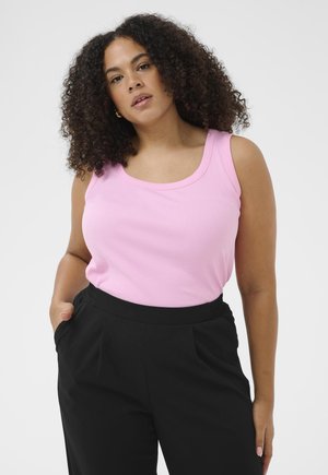 Frau mit lockigem Haar, die ein pinkfarbenes ärmelloses Oberteil und schwarze Hose trägt, posiert mit einer Hand in der Tasche vor einem schlichten Hintergrund.