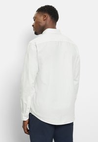 Camicia bianca a maniche lunghe con colletto button-down e spacchi laterali, caratterizzata da una texture liscia e un design aderente. Abbinata a pantaloni scuri.