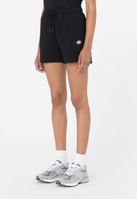 Pantalones cortos de algodón negros con cintura elástica y cordón de ajuste, que cuentan con bolsillos laterales y un parche de logo colorido. Combinados con zapatillas grises.