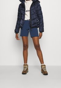 Tmavě modrá prošívaná bunda s kapucí, zdobená geometrickým vzorem. Kombinována s modrými šortkami a hnědými turistickými botami s béžovými tkaničkami.