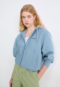 Sudadera de cremallera azul claro con un bolsillo frontal, que presenta un logo bordado. Combinada con pantalones verdes texturizados, que exhiben un corte relajado.