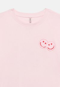 Ανοιχτό ροζ κοντομάνικο μπλουζάκι με λαιμόκοψη crew, με δύο επικαλυπτόμενα κόκκινα γραμματόσημα χαμογελαστού προσώπου στην αριστερή πάνω πλευρά του στήθους.