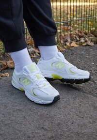 Vita idrottsskor i vitt med meshmaterial och gult-gröna accenter, utrustade med en dämpad sula och snörningsdesign, visade på grå asfalt.