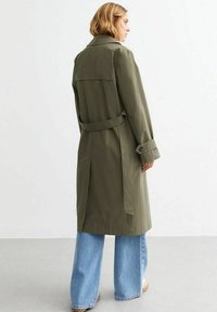 Olivgrön trenchcoat med bälte i midjan, axelklaffar och knäppta manschetter. Bärs över ljusblå vida jeans.