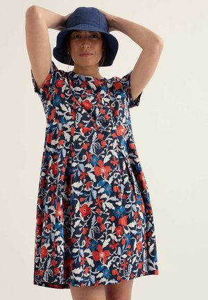 Femeie purtând o pălărie tip bucket de culoare bleumarin și o rochie florală până la genunchi, cu flori roșii, albastre și albe, pe un fundal simplu.