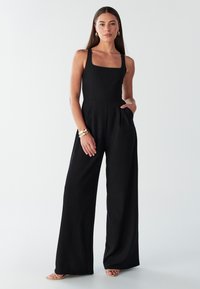 Black jumpsuit with a square neckline, fitted bodice, wide legs, and side pockets. Smooth fabric with a subtle sheen. Accessories include a gold bracelet.