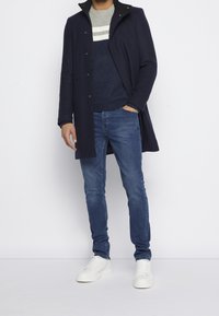 Marineblauer maßgeschneiderter Mantel mit hohem Kragen, grau-blau gestreifter Strickpullover, blaue Slim-Fit-Jeans und weiße Sneakers.