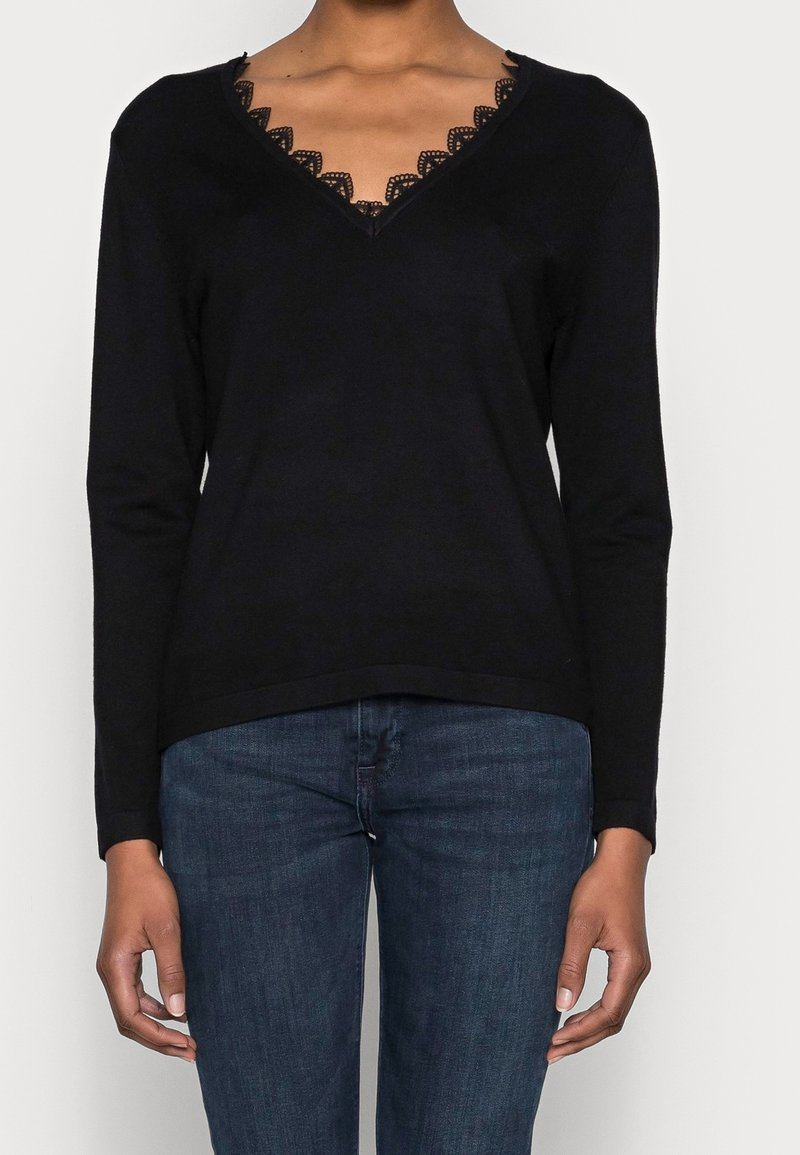 Pull noir à manches longues avec col en V et bordure en dentelle porté avec un jean denim bleu sur une personne devant un fond uni.