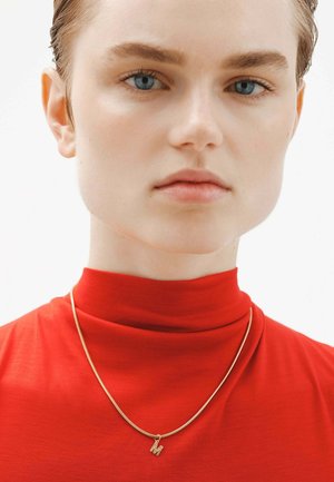 Collar de oro con un pequeño colgante en forma de la letra "M," llevado sobre una blusa roja de cuello alto. Textura de tela suave y un contraste de color brillante.