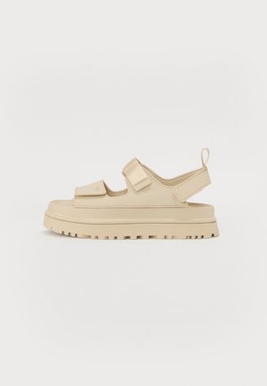 Beige sandal med tyk sål, to justerbare remme over foden og en hælrem med trækflig, vist på hvid baggrund.