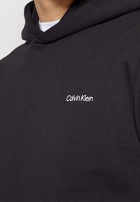 Zwarte hoodie gemaakt van zacht materiaal met een wit "Calvin Klein" logo op de borst. Eenvoudig en minimalistisch ontwerp.