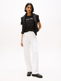 T-shirt noir avec le logo "Tommy Hilfiger" en blanc, associé à un jean blanc taille haute et des mocassins noirs. Un pull est drapé sur les épaules.