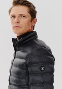 Czarna kurtka puchowa z wysokim kołnierzem, zamkiem błyskawicznym i pikowaną teksturą. Posiada okrągłą łatę z logo na lewym rękawie.