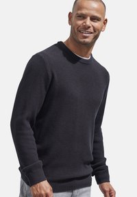 Dunkel gestrickter Pullover mit einem strukturierten Muster, rundem Ausschnitt und gerippten Bündchen sowie Saum. Getragen über hellen Jeans.