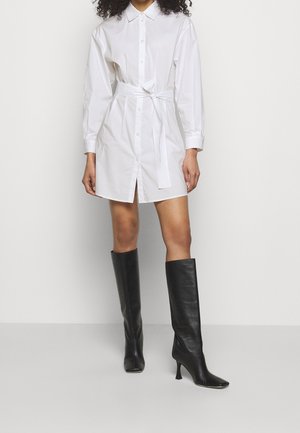 Femme portant une robe chemise blanche à boutons avec une ceinture à la taille, associée à des bottes noires à talons hautes jusqu'aux genoux, sur un fond uni.