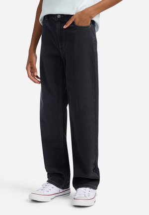 Personne portant un jean noir ample et des baskets blanches, avec une main dans la poche et un haut de couleur claire partiellement visible.