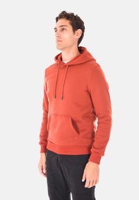 Uomo in piedi, che indossa un felpa color ruggine con tasca frontale e pantaloni neri, che guarda leggermente a sinistra su uno sfondo semplice.