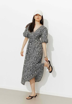 Femme en robe midi à fleurs noire et blanche, casquette blanche, boucles d'oreilles créoles et sandales à talons noires, ajustant une chaussure contre un mur blanc.