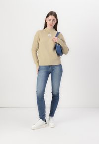 Beżowy sweter z prążkowanego materiału z wysokim kołnierzem, niebieska torebka i jasnoniebieskie dżinsy. Model ma na sobie białe trampki z czarnymi akcentami.
