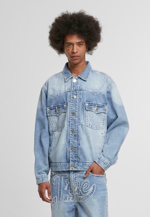 Junger Mann mit Afrofrisur, der eine hellblaue Jeansjacke und Jeans trägt, auf denen das Logo "True Religion" gestickt ist.