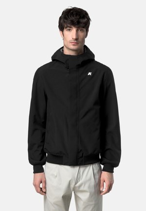 Giovane uomo che indossa una giacca con cappuccio nera con logo sul petto e pantaloni di colore chiaro, in piedi davanti a uno sfondo uniforme.