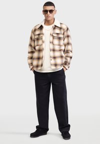 Braun- und cremefarbener karierter Flanelljacke mit weichem Kragen, über einem hellen Hemd getragen, kombiniert mit schwarzen Hosen und dunklen Schuhen.