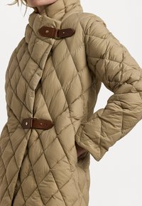 Casaco puffer acolchoado em bege claro com um colarinho alto, apresentando costura diagonal, alças em pele castanha e um corte descontraído.