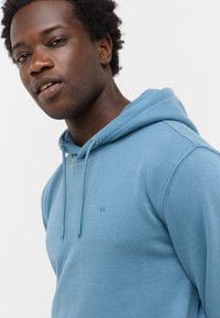 Młody mężczyzna w jasnoniebieskiej bluzie z kapturem, zwrócony nieco w lewo z neutralnym wyrazem twarzy, na jednolitym białym tle.