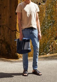 Beige bomulls-t-shirt, ljusblå slitna jeans, svarta remsandaler och en flerfärgad denimväska med handtag.